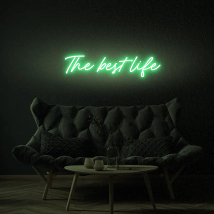 The Best Life LED Neon Light Sign in green, featuring script lettering illuminated above a dark tufted sofa.