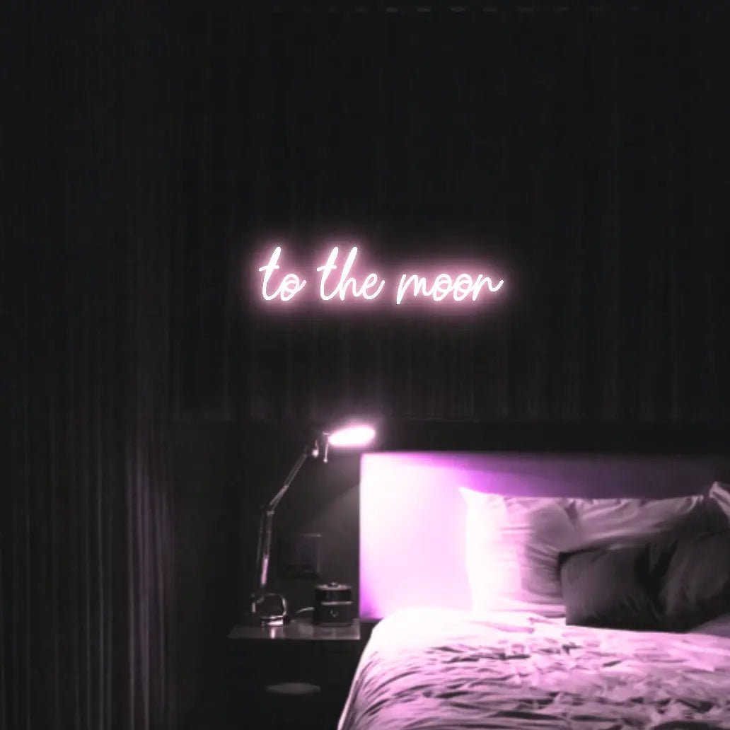 To The Moon LED Neon Light Sign in pale pink, featuring script lettering illuminated above a bed in a dark bedroom.